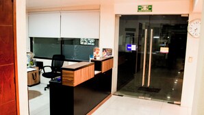Un mostrador de recepción con superficie de madera y base negra, una silla de oficina negra, y una puerta de cristal que da a un pasillo.