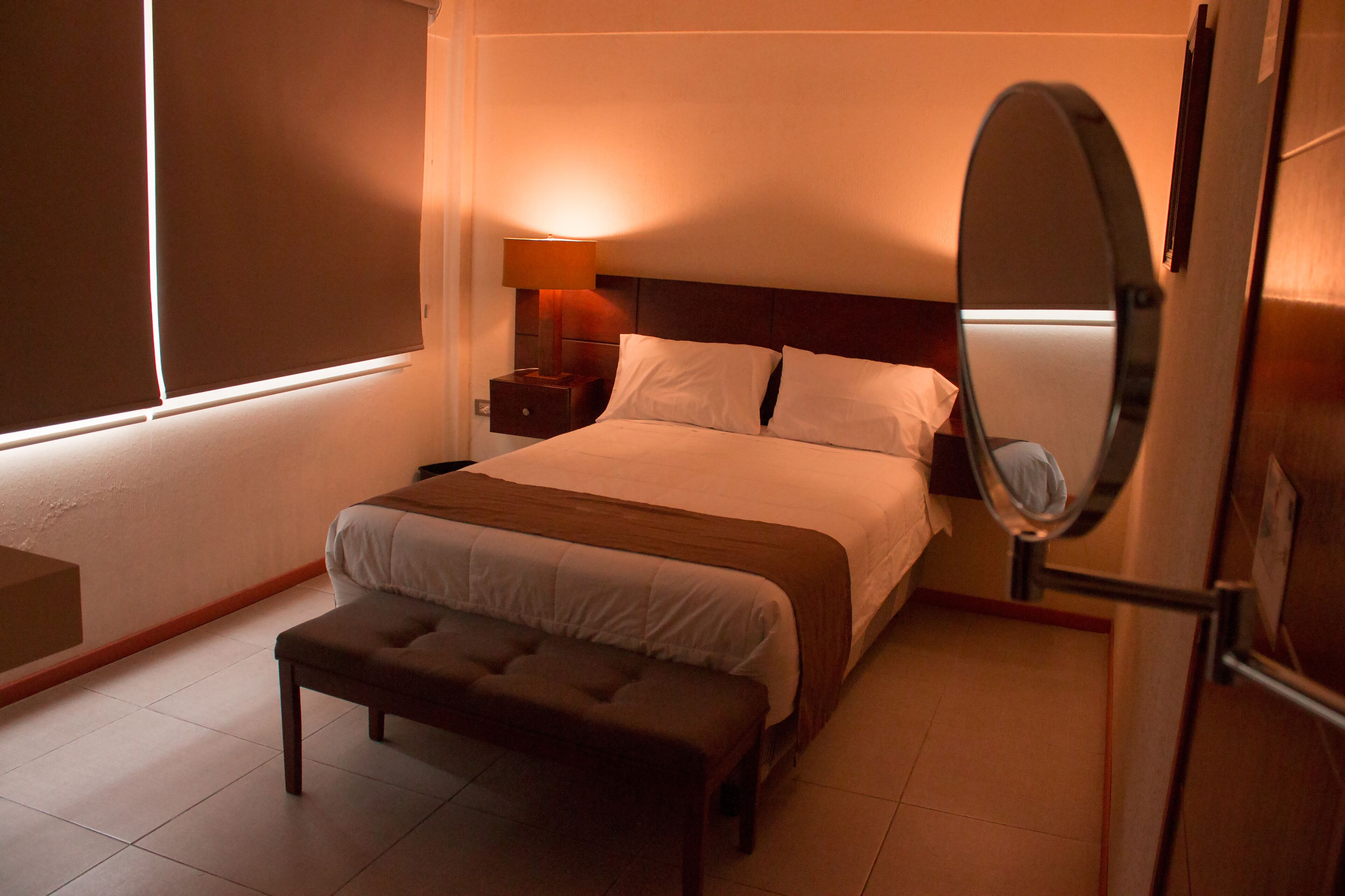 Habitación de hotel con una cama grande, mesita de noche con lámpara, un banco y un espejo.