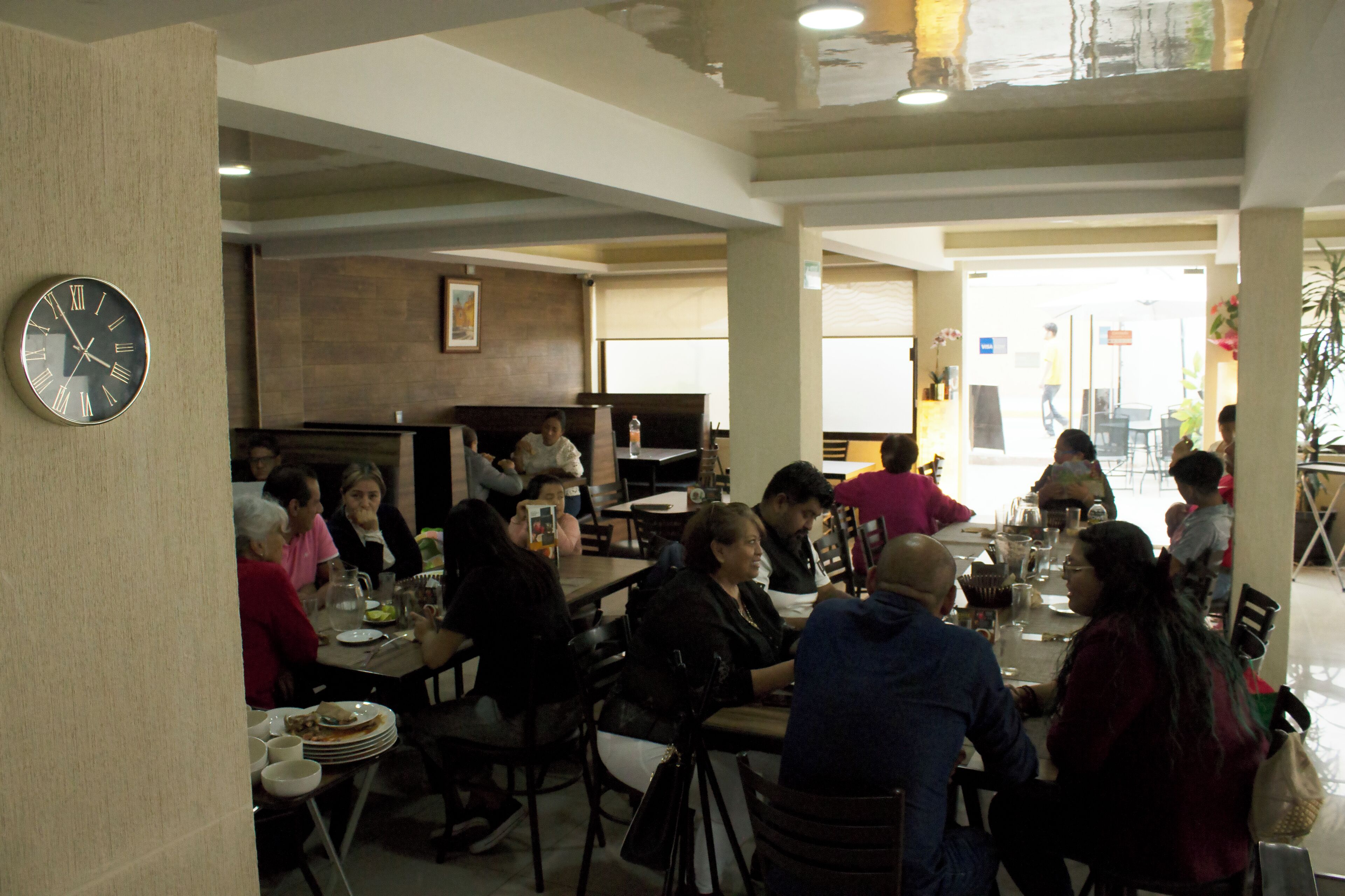A restaurant with multiple tables and chairs, a clock on the wall, and a few people dining.