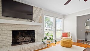 Una sala de estar con chimenea, un televisor de pantalla plana montado encima, un sillón cómodo y un ventanal con persianas.