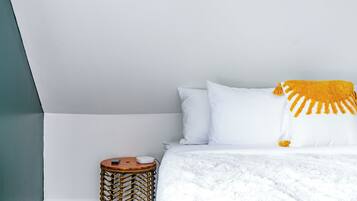 Un dormitorio con cama, almohadas blancas, una colcha blanca, una mesita de noche redonda de mimbre y una manta amarilla.