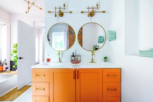 A bathroom with two round mirrors, an orange vanity, a white sink, and a door leading to another room.