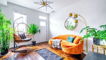 Een moderne woonkamer met een houten vloer, een hangstoel, een oranje bank en een ronde spiegel met een plant.