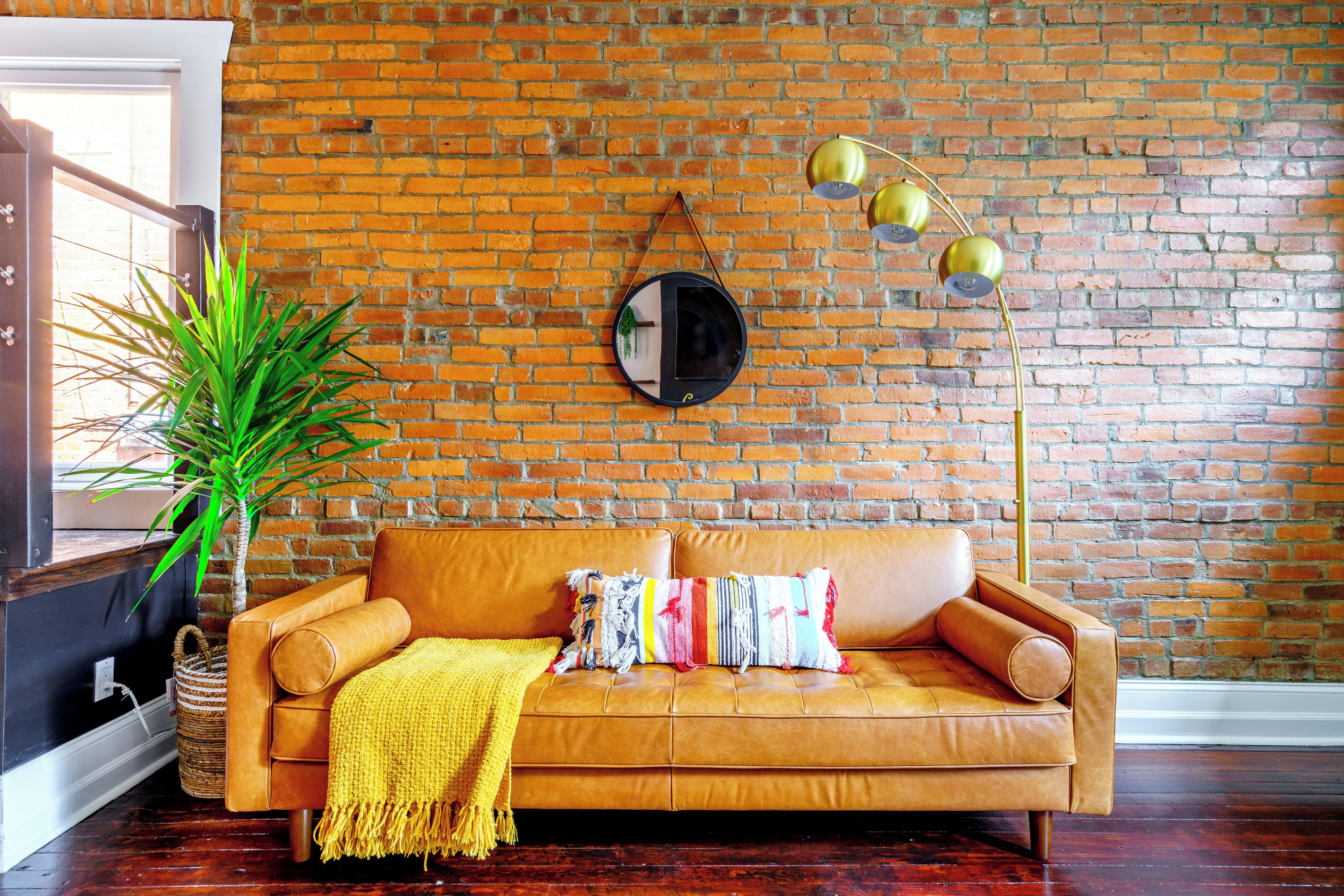 A room with a brick wall, a leather sofa with cushions, a floor lamp, and a potted plant.