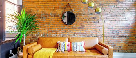 A room with a brick wall, a leather sofa with cushions, a floor lamp, and a potted plant.