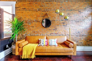 A room with a brick wall, a leather sofa with cushions, a floor lamp, and a potted plant.