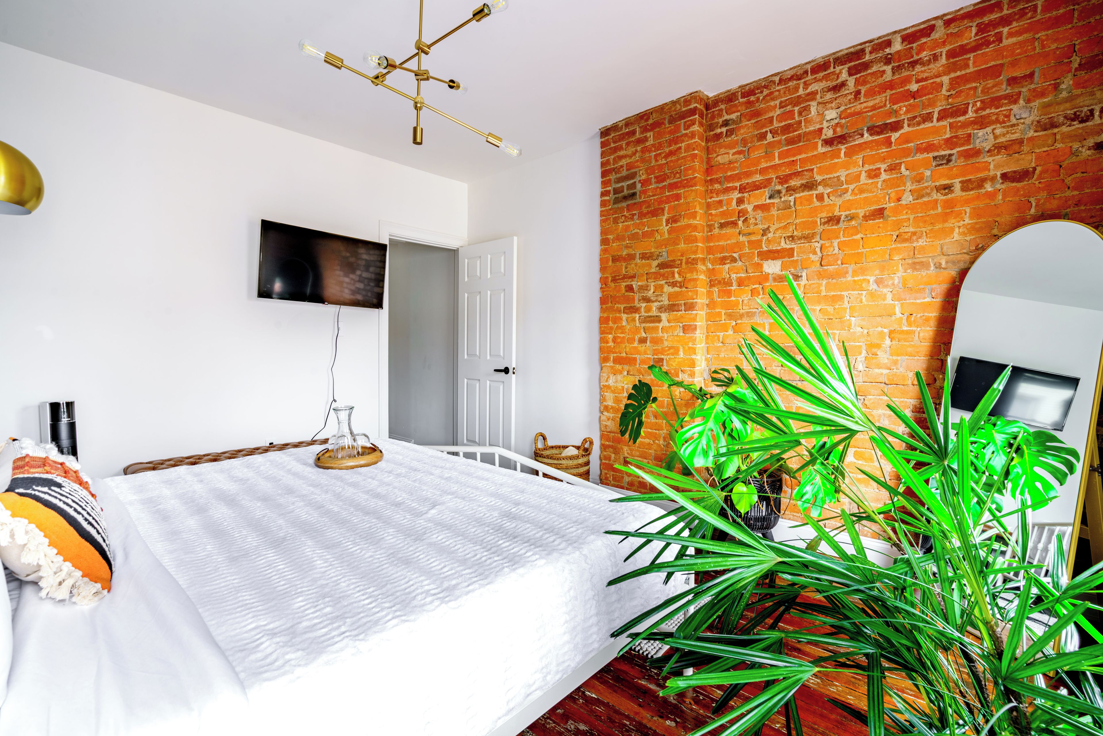 Un dormitorio con pared de ladrillo, cama con cubrecama blanco, planta grande y televisor montado en la pared.