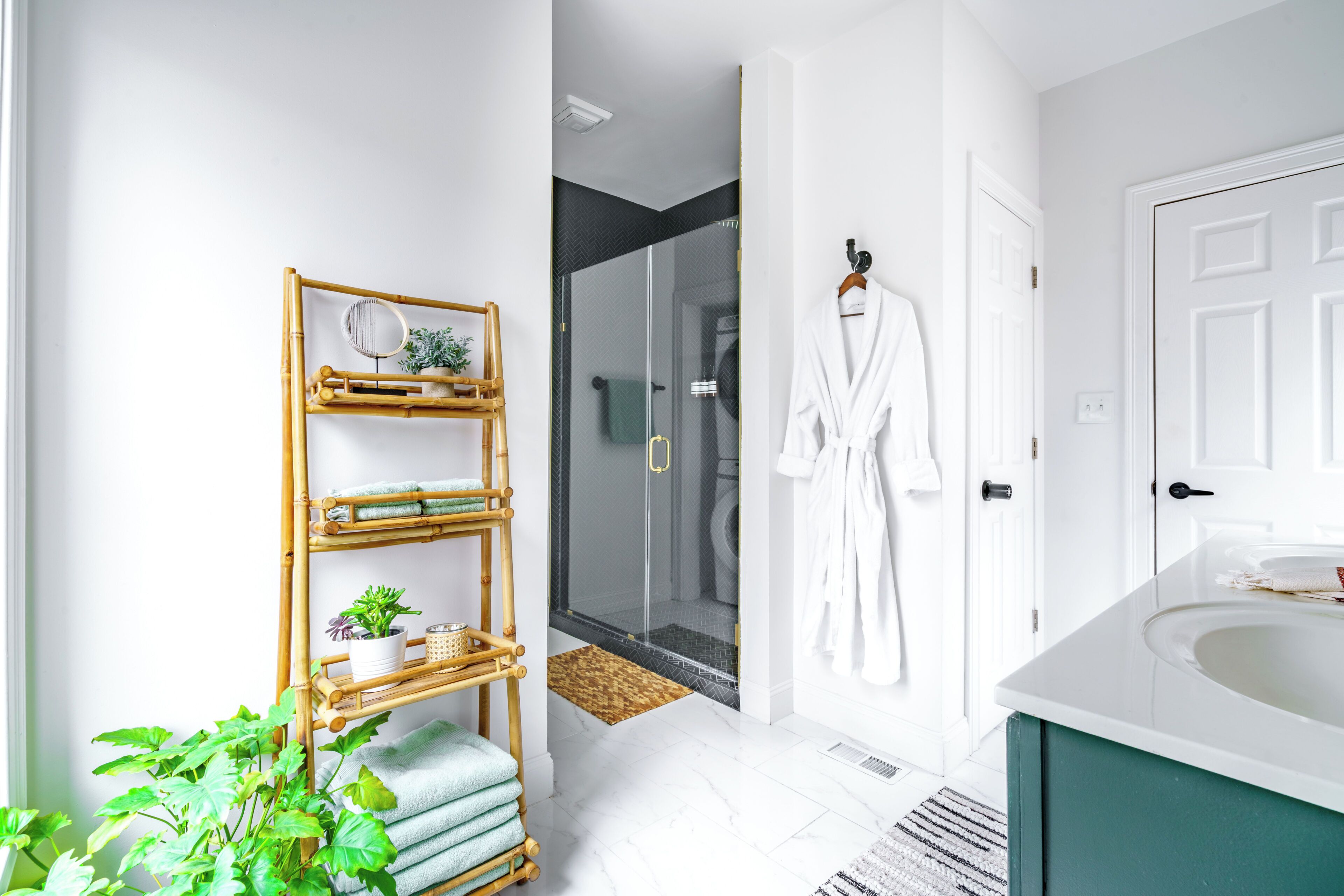 A bathroom with a white bathtub, a bamboo shelf with towels and a plant, a glass shower enclosure, and a white robe hanging on the wall.
