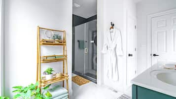 A bathroom with a white bathtub, a bamboo shelf with towels and a plant, a glass shower enclosure, and a white robe hanging on the wall.