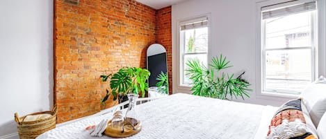 Een kamer met een bakstenen muur, een bed met witte lakens, een raam met zonwering en een plant.