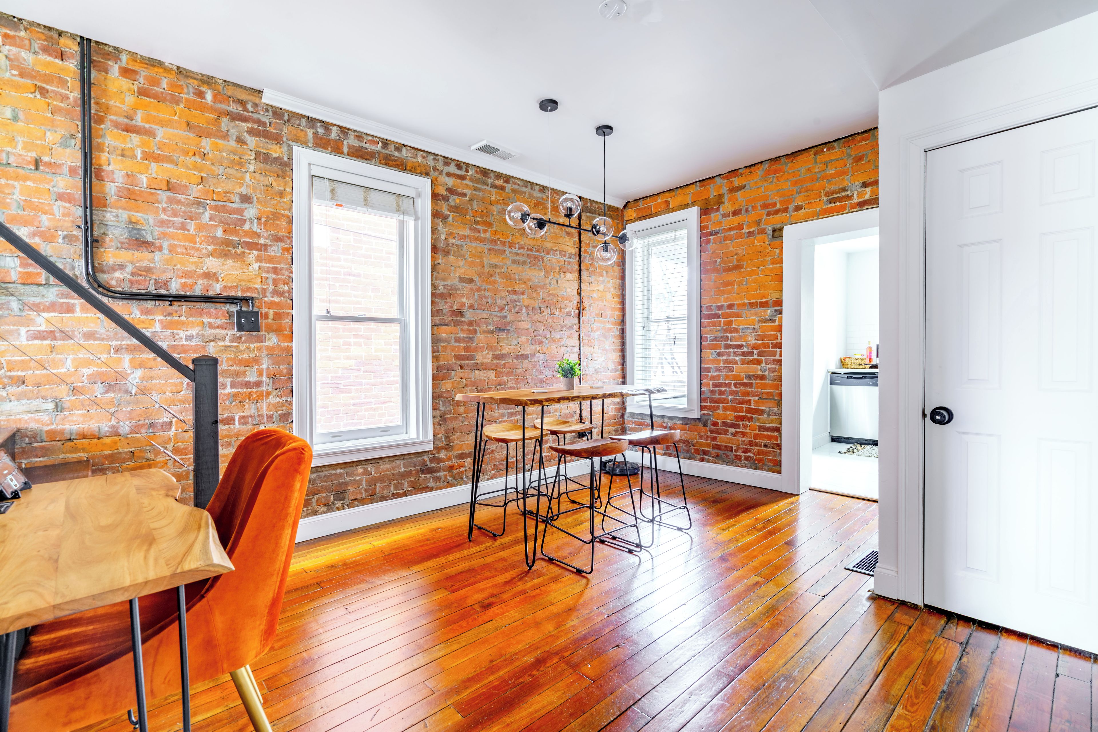 A room with a brick wall, wooden floor, a dining table with chairs, a hanging light fixture, and a white door.