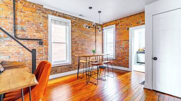 A room with a brick wall, wooden floor, a dining table with chairs, a hanging light fixture, and a white door.
