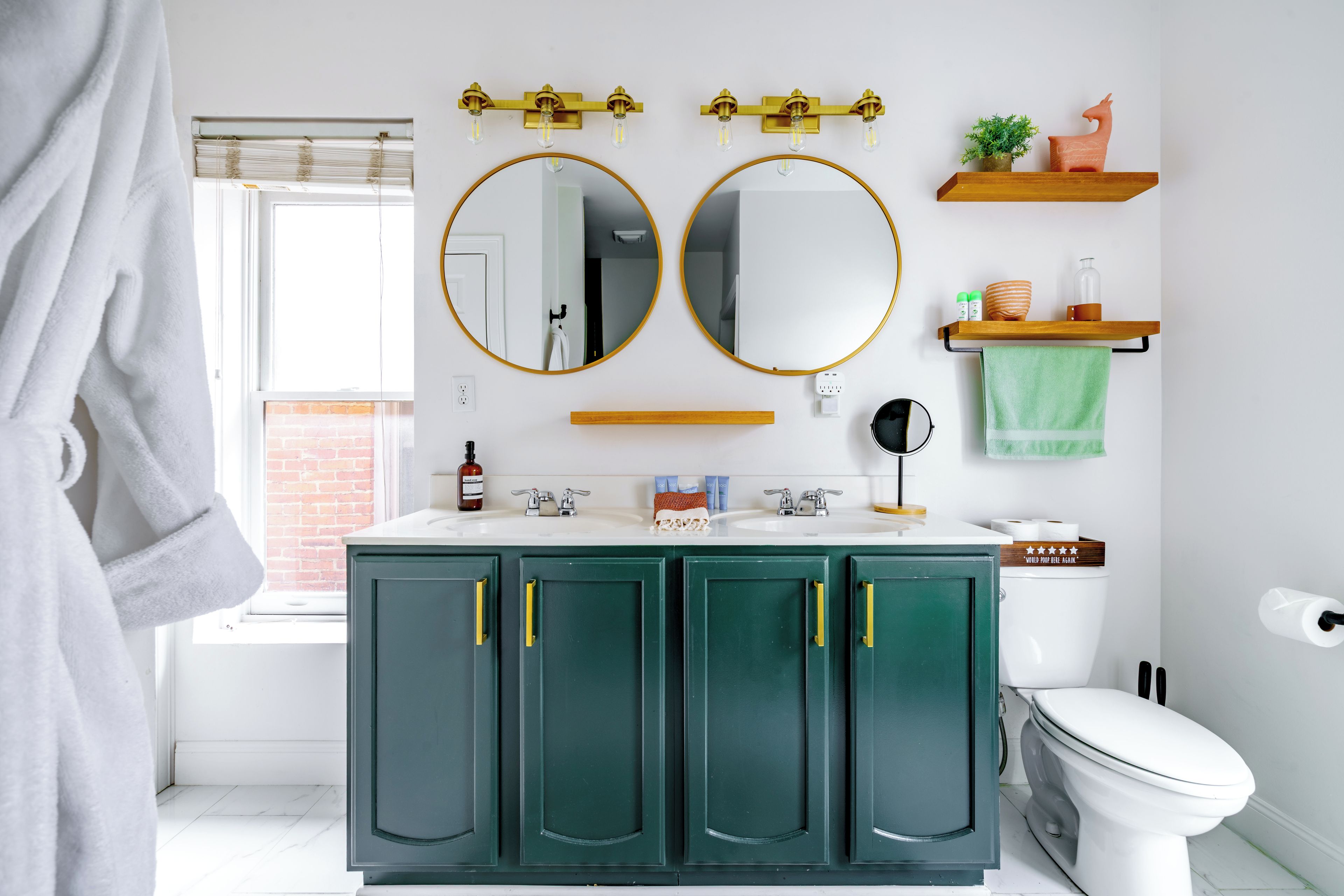 Une salle de bain avec des toilettes blanches, des meubles verts, deux miroirs ronds et une fenêtre avec des stores.