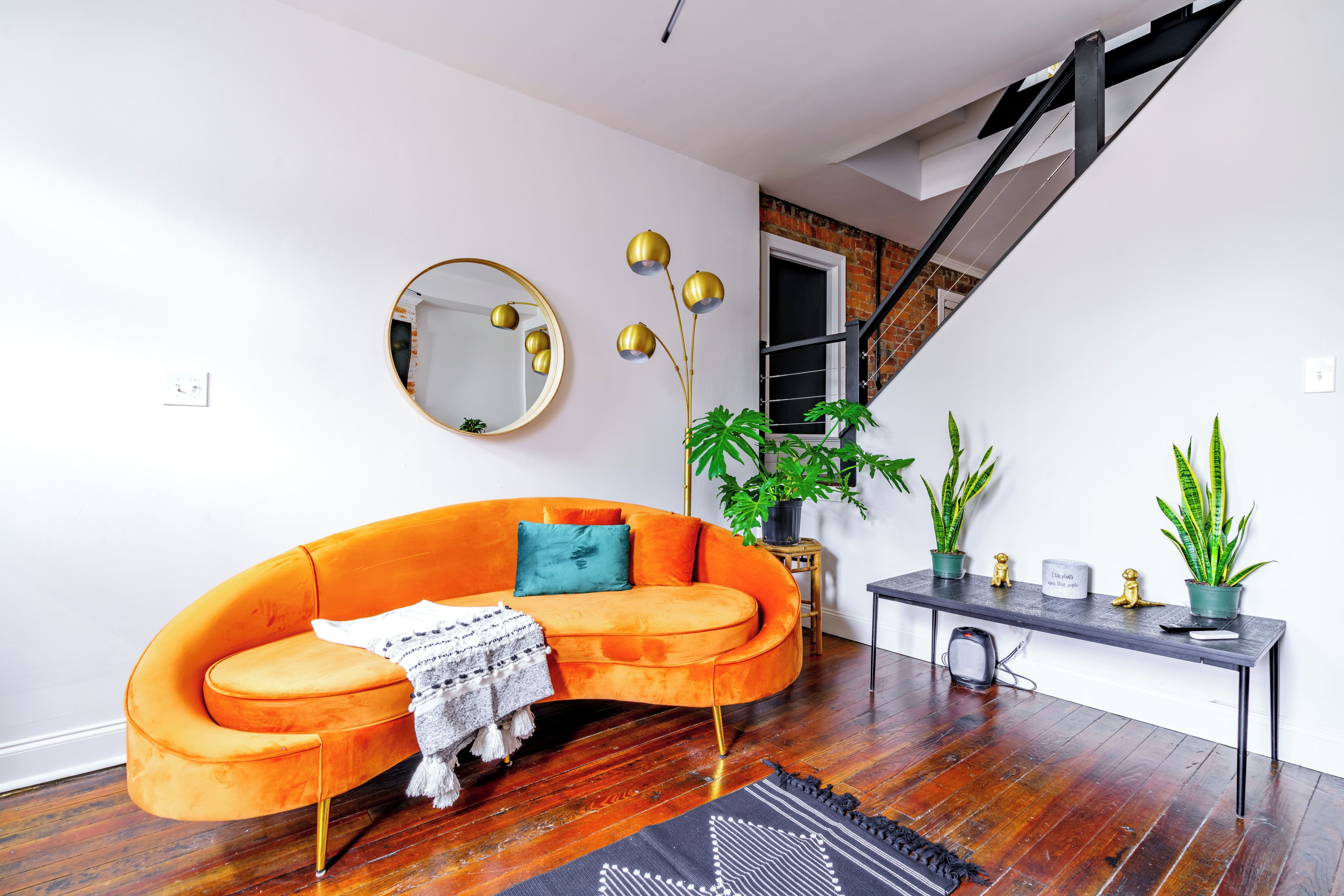 A modern living room with a curved orange sofa, a round mirror, a console table with plants, and a staircase.