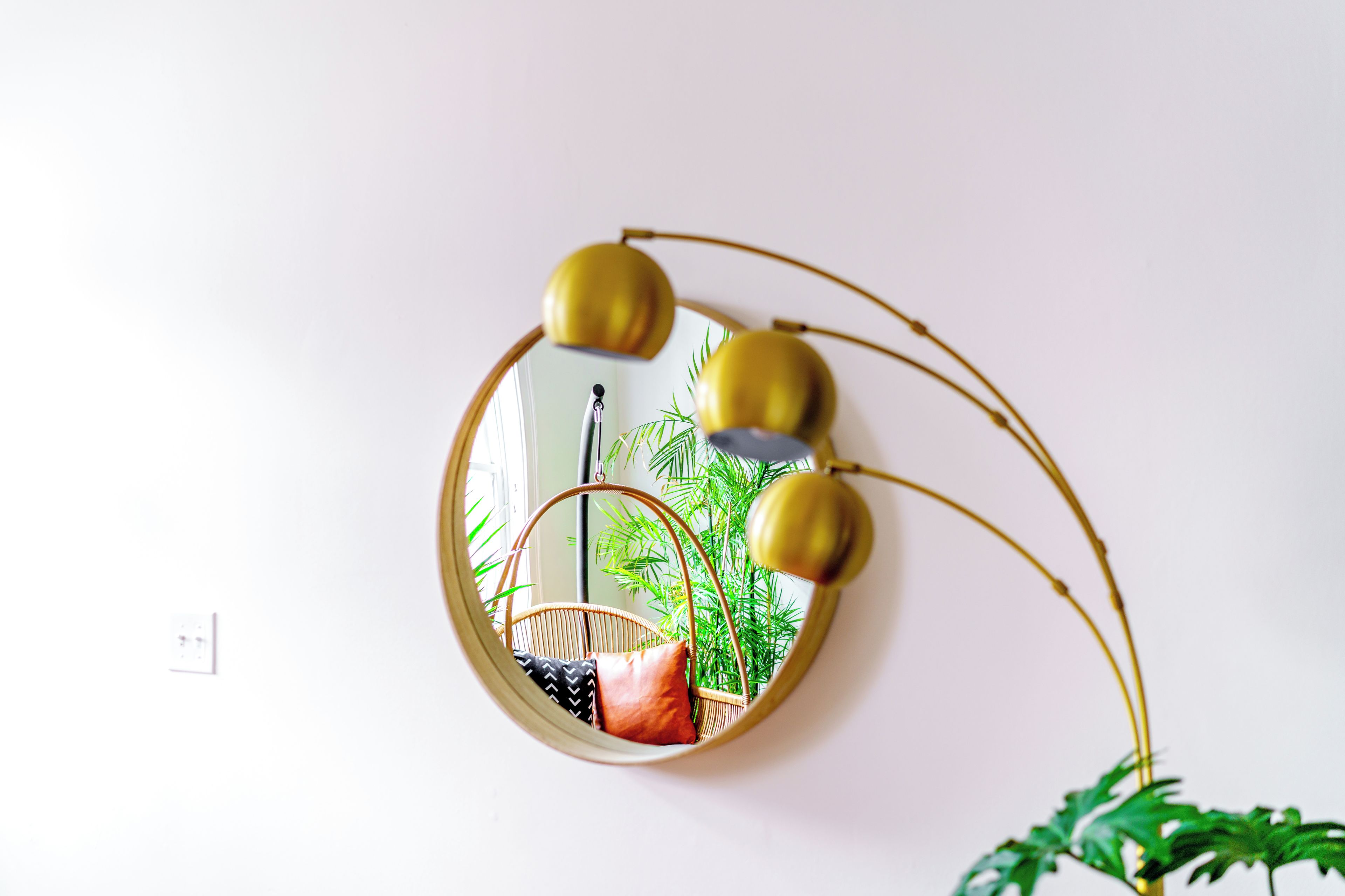 A round mirror with a gold frame and three gold spotlights, reflecting a wicker chair and a plant.