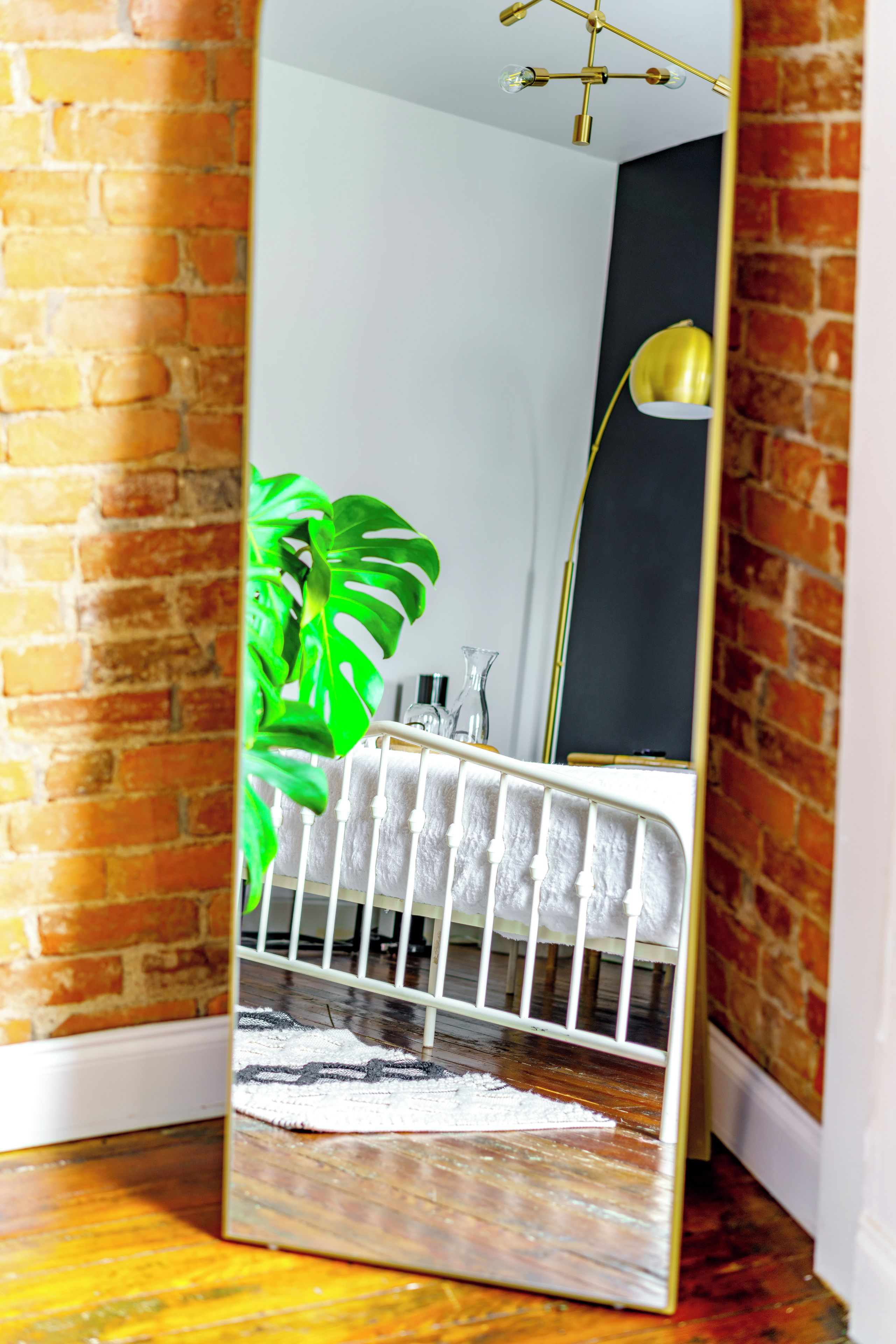 A mirror reflecting a room with a brick wall, a white radiator, a green plant, and a white baby crib.