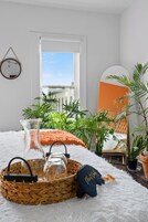 A room with a white furry rug, a woven basket with glass bottles and a black object, green plants, and a large mirror with an orange frame.