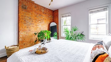 Een kamer met een bakstenen muur, een bed met witte lakens, een raam met zonwering en een plant.