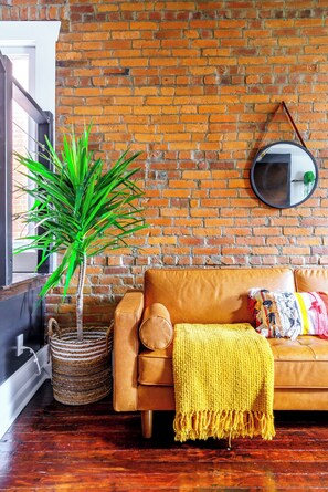 A room with a brick wall, a round mirror, a brown leather sofa, a yellow knitted throw, and a green plant in a woven basket. - Jungle House Urban Lofts - Short North & Convention Center (Columbus)