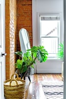 A room with a brick wall, a white door, a window, a mirror leaning against the wall, and a potted plant.