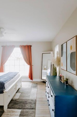 Une chambre à coucher avec un lit, une commode, une fenêtre avec des rideaux et des œuvres d’art murales.