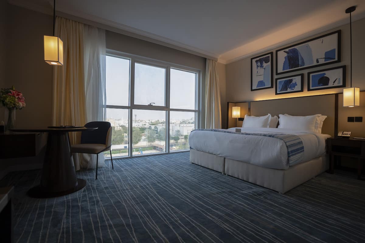 A hotel room with a large bed, a desk, a chair, a view through the window, and framed pictures on the wall.