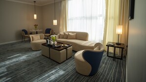 Una habitación de hotel moderna con un sofá color beige, un sillón azul, una mesita redonda con un florero y una mesa de centro.