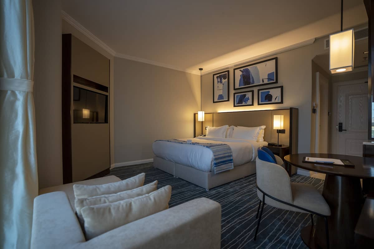 A hotel room with a bed, bedside tables, a desk, a chair, and wall-mounted artwork.