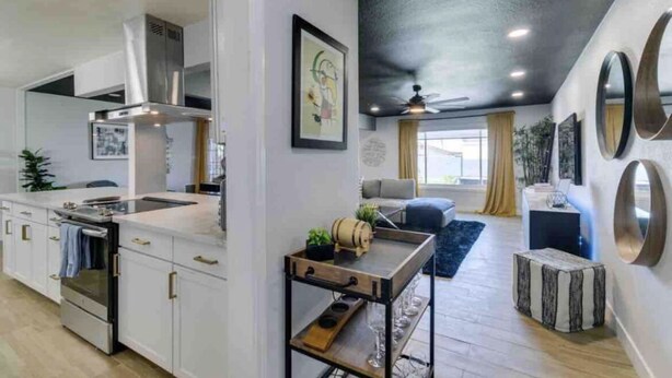 Een moderne keuken met witte kasten, roestvrijstalen apparatuur en een centraal eiland. Er is een zithoek met een bank en salontafel. De kamer heeft een zwart plafond en een groot raam met gordijnen.