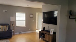 A living room with a dark-colored sofa, a wooden entertainment unit, a flat-screen TV, and a window with blinds. - Amazing 3 bedroom home, perfect for families! (Glendale)