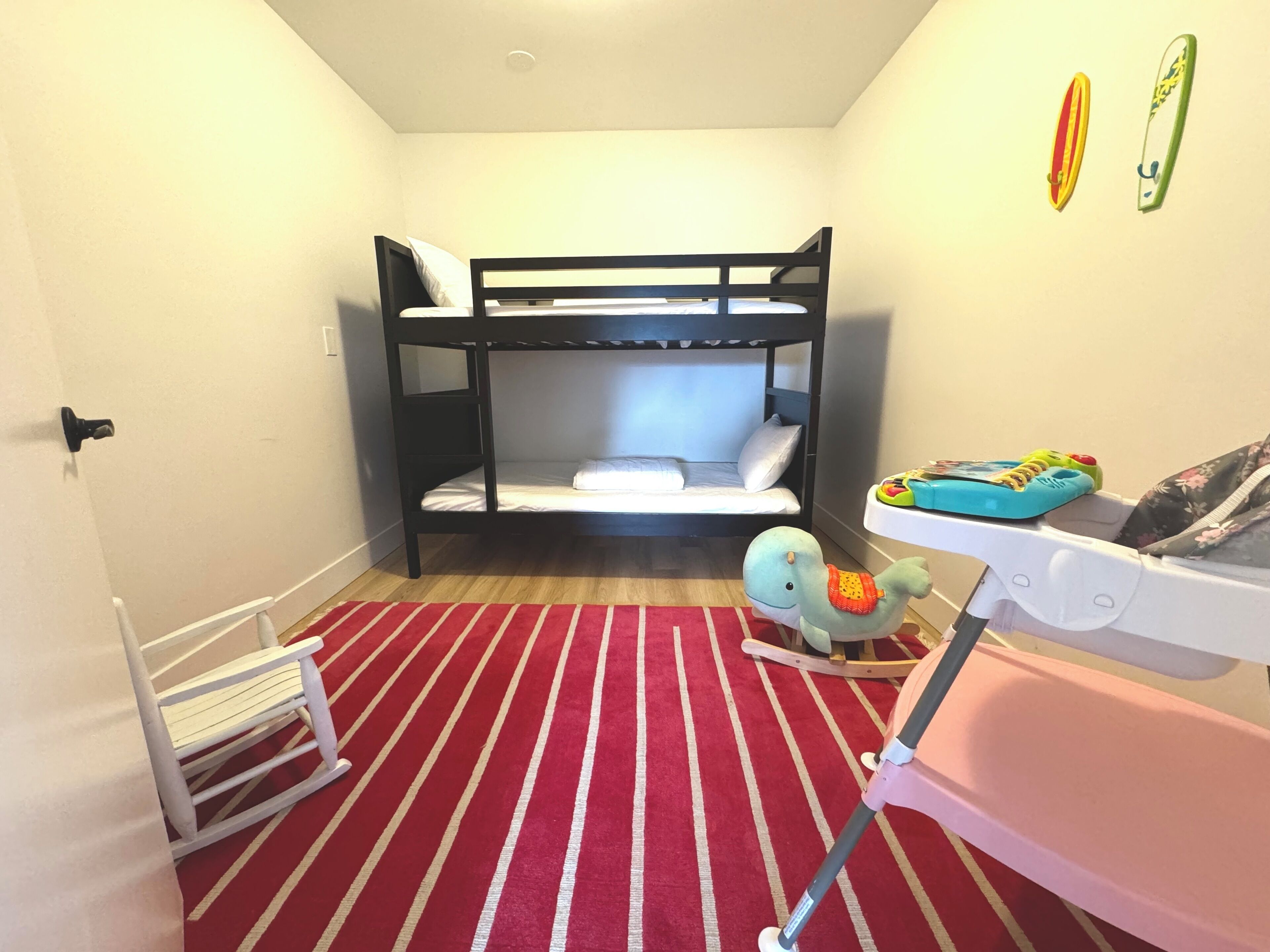 Et værelse med køjeseng, et rødt stribet tæppe, en lille hvid hylde og en babystol.