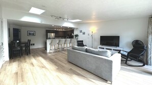Un salon moderne avec un canapé gris, un plancher en bois, un coin repas avec une table et des chaises, une cuisine avec des meubles sombres et un téléviseur posé sur un meuble.