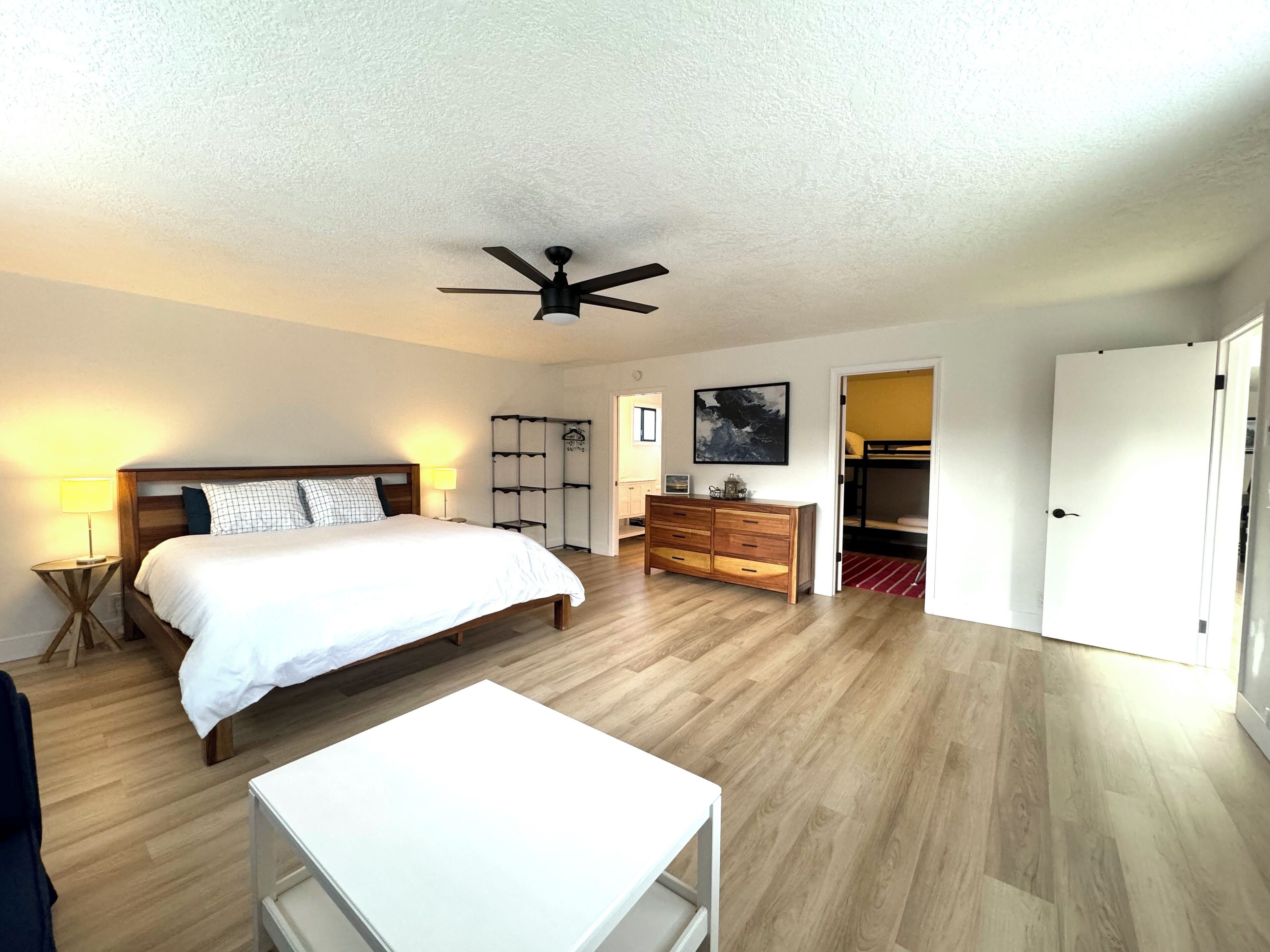 A bedroom with a bed, a desk, a wardrobe, a ceiling fan, and a door leading to another room.