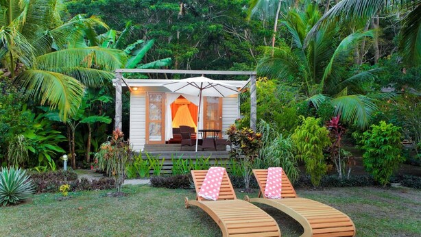 Un jardín tropical con una terraza de madera, dos sillones de relax con toallas rosas y una zona de estar cubierta con una mesa y sillas.