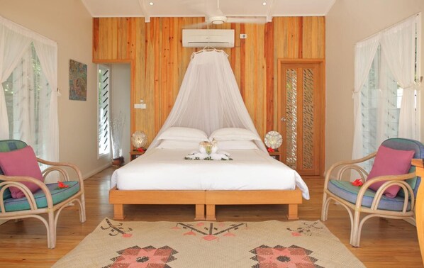 A bedroom with a wooden paneled wall, a canopy bed, two wicker chairs, a decorative rug, and a bedside table with a lamp. - Deluxe Oceanfront Villa with indoor and outdoor private shower and private deck. (Somosomo)