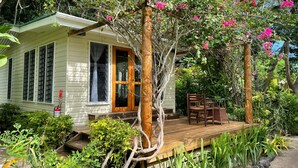 Una casita con terraza de madera, rodeada de abundante vegetación y plantas con flores.