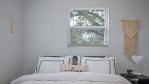 A bedroom with a bed, a bedside table with a lamp, a macrame wall hanging, and a window with blinds. - Boho Villa B (Tampa)