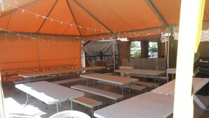 Een overdekt eethoekje buiten met meerdere tafels en banken, een bakstenen muur en een houten omheining op de achtergrond.