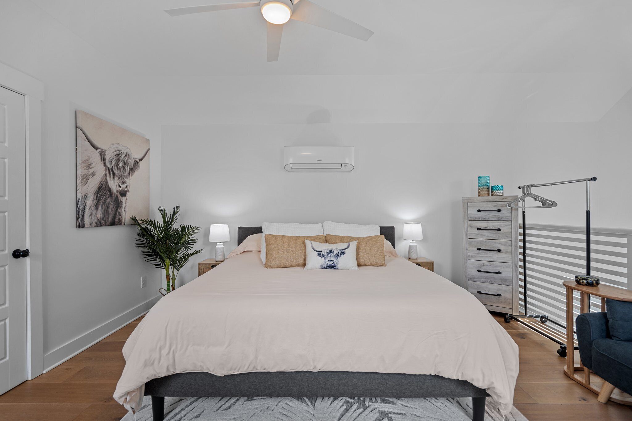 Une chambre à coucher comprenant un lit, des lampes de chevet, un ventilateur de plafond, un climatiseur mural, une plante, une commode et une fenêtre avec des stores.