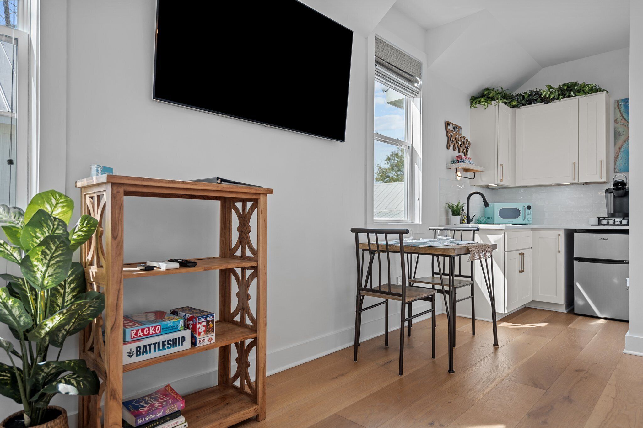 A modern kitchen with white cabinets, a wooden dining table, and a large flat-screen TV mounted on the wall.