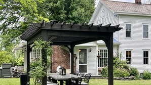 Un patio cubierto con una mesa y sillas, un muro de ladrillo y una casa blanca al fondo.