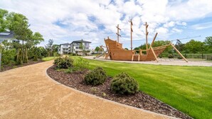 A wooden ship-shaped playground in a well-maintained park with a paved path, surrounded by greenery and residential buildings in the background. - Haffhaus 1 - Haffhaus (Garz)
