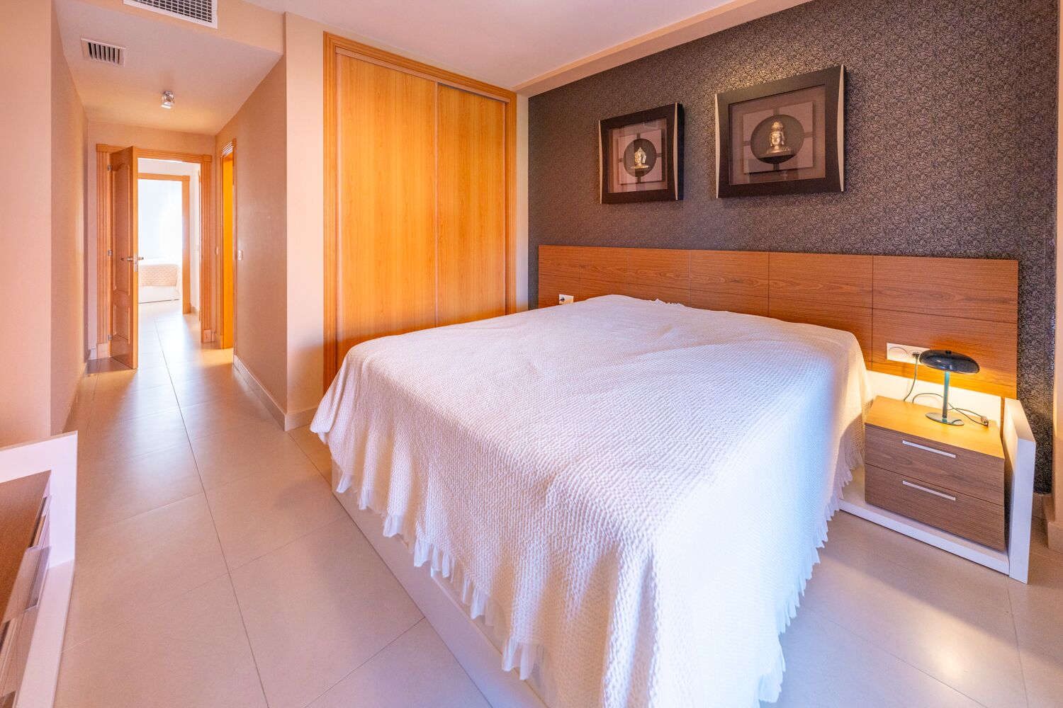 Un cuarto de hotel con una cama grande, un cabecero de madera, una mesita de noche con lámpara y dos cuadros enmarcados en la pared.