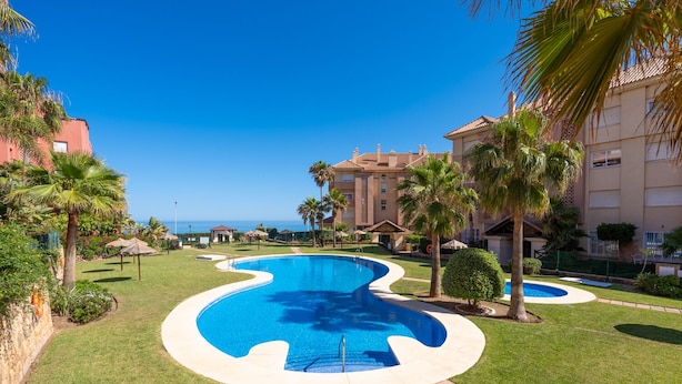 Ein Pool mit geschwungener Kante, umgeben von gepflegtem Rasen und Palmen, mit Gebäuden im Hintergrund.
