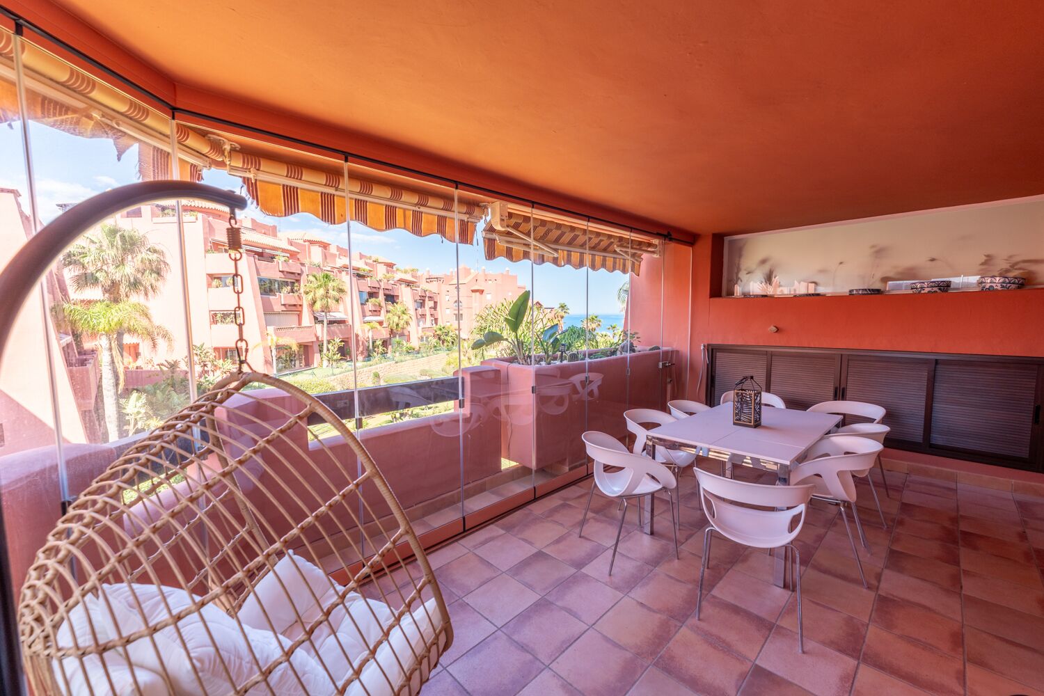 Un balcón con una silla de mimbre, una mesa con sillas y vistas a una zona residencial.