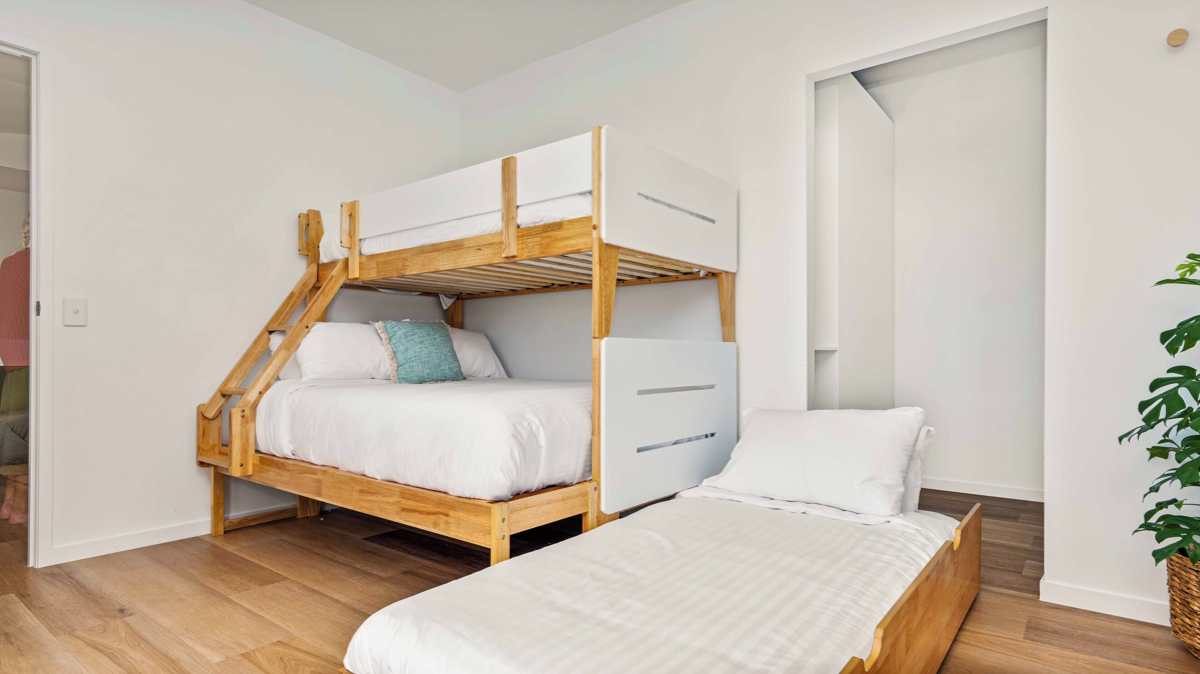 Un lit superposé avec une structure en bois et du linge de lit blanc, un lit simple avec du linge de lit blanc, et une plante en pot dans un panier.