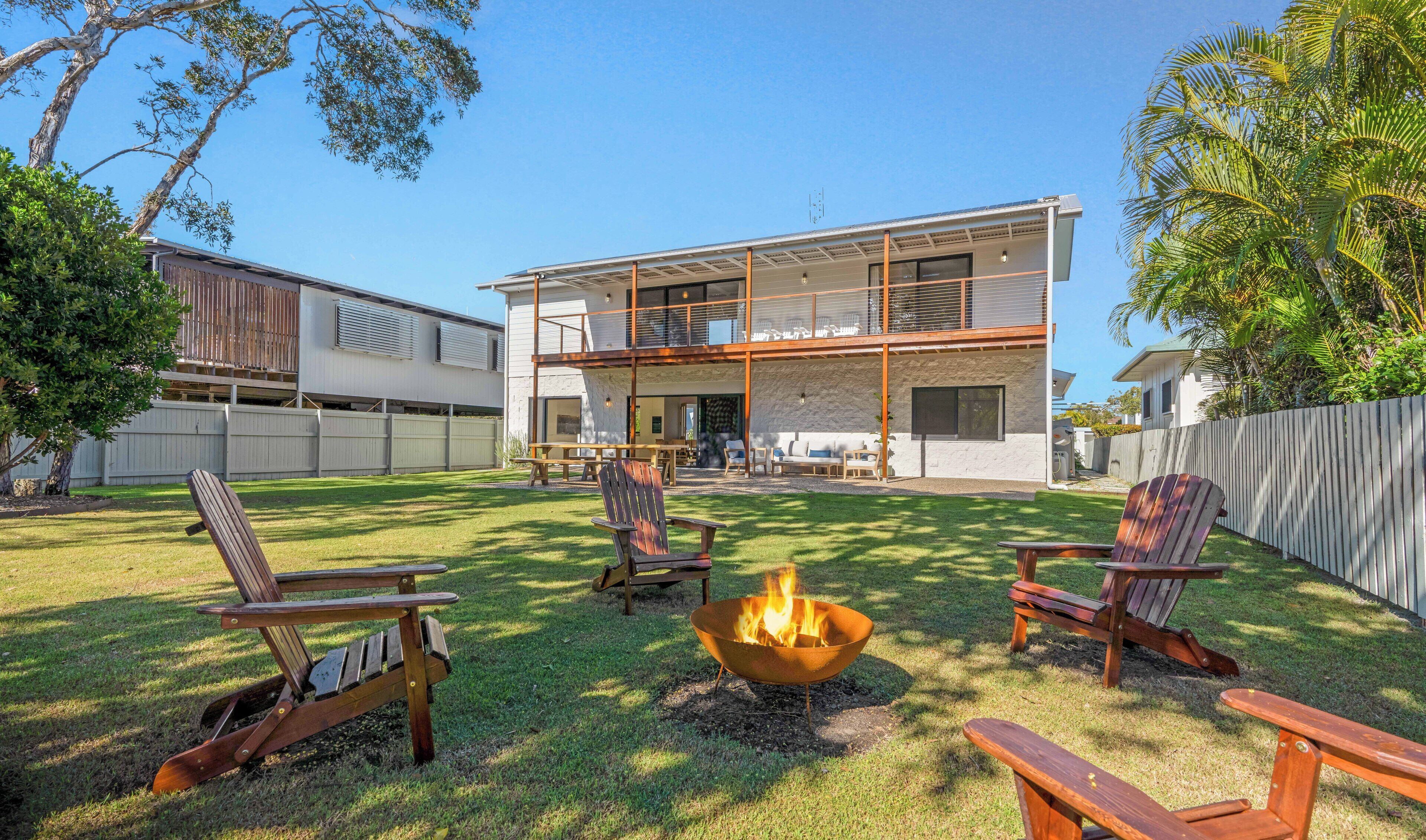 A modern two-story house with a backyard featuring a fire pit, wooden chairs, and a dining area.
