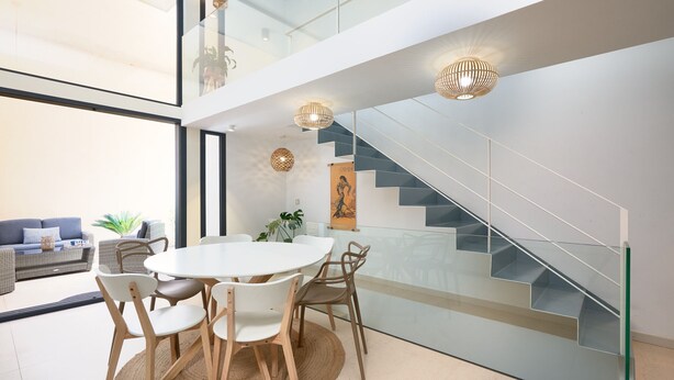 Um interior moderno com uma escada de vidro, uma sala de jantar com uma mesa redonda e cadeiras, e um sofá ao fundo.
