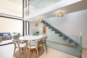Un interno moderno con una scala in vetro, una zona pranzo con un tavolo e sedie rotonde, e un divano sullo sfondo.