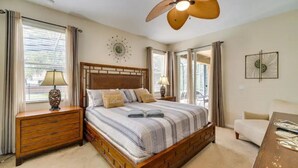 A bedroom with a wooden bed, two bedside tables with lamps, a ceiling fan, and a view of the outdoors through large windows.